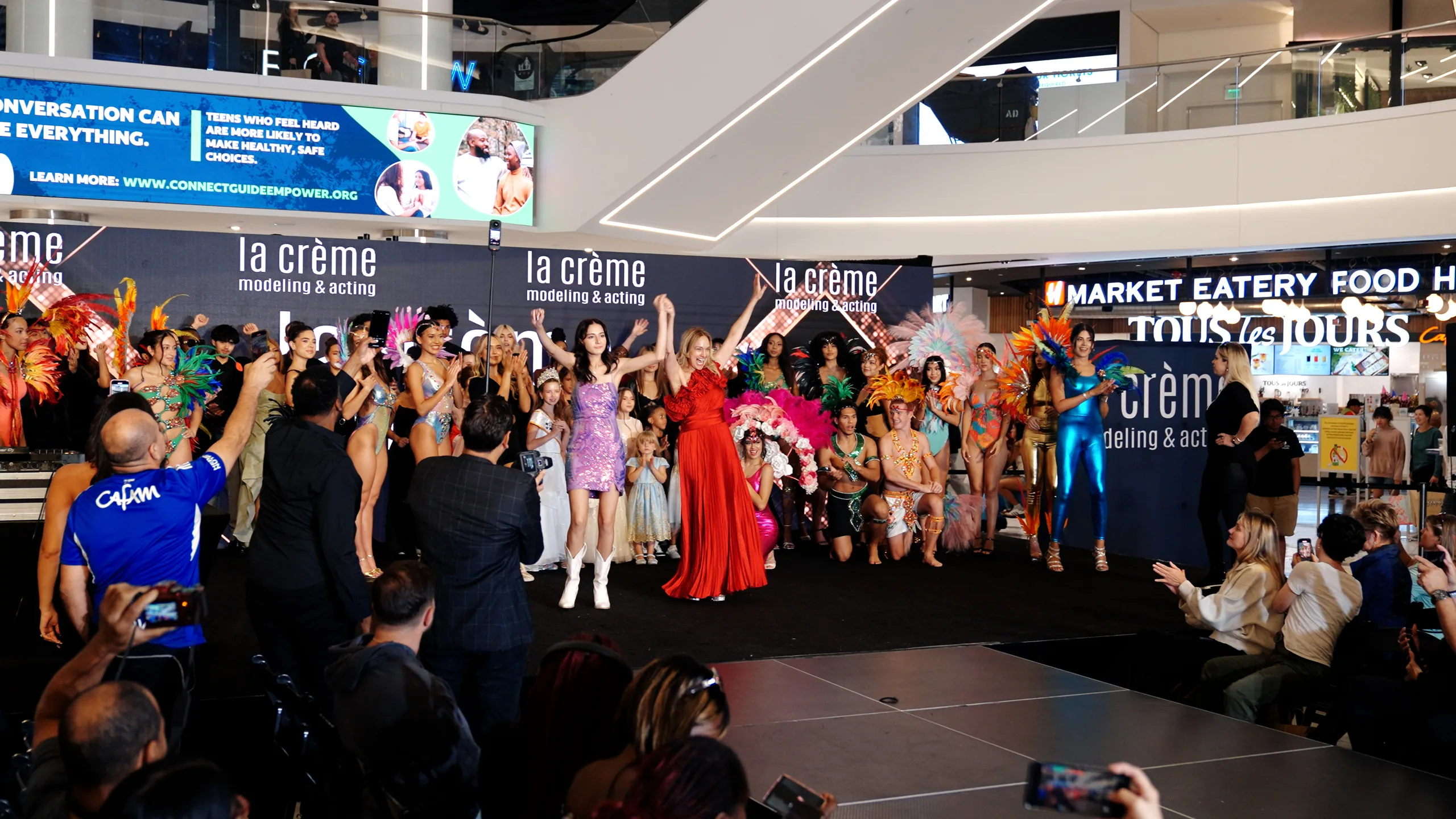 LaCreme fashion show finale at American Dream Mall — models in feathered showpieces and the founders with arms raised in front of the la crème modeling & acting backdrop.